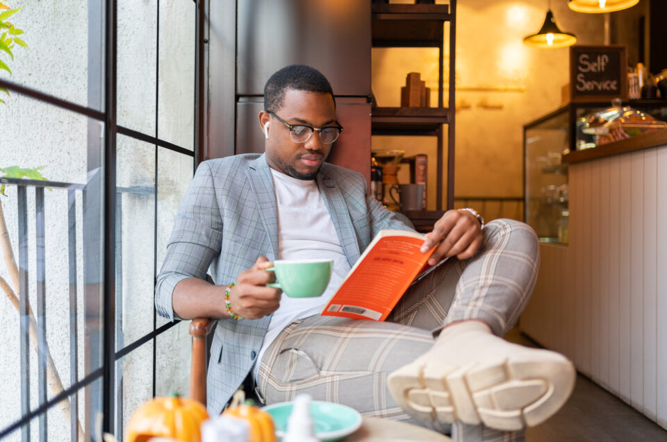 A Young Man Reading Book And Listening Music On Smartphone At Coffee Shop